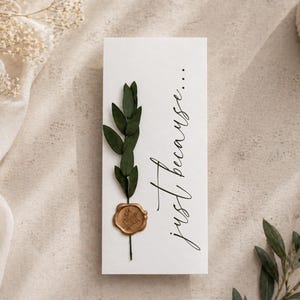 Puede incluir: Una tarjeta rectangular blanca con las palabras "just because..." en escritura elegante. Una ramita de hojas verdes y un sello de cera dorado adornan la tarjeta. La tarjeta está sobre una superficie de tonos neutros, con acentos florales.