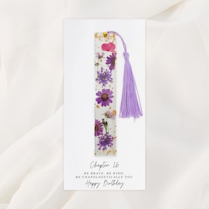 Puede incluir: Un marcapáginas rectangular con flores moradas y rosas incrustadas, un acento dorado y una borla lavanda. El marcapáginas está en una tarjeta blanca con el texto "Chapter 16, Be brave, be kind, be unapologetically you, Happy Birthday."