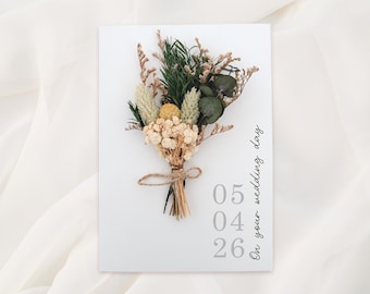 May include: A white card with a dried flower bouquet tied with twine. The bouquet includes green, beige, and yellow flowers. The card has the text "On your wedding day" and the date "05 04 26" in gray.