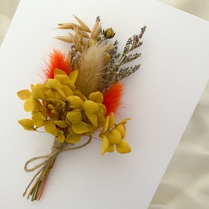 May include: A small bouquet of dried flowers, featuring yellow hydrangeas, orange feathers, and dried grasses, tied together with twine.