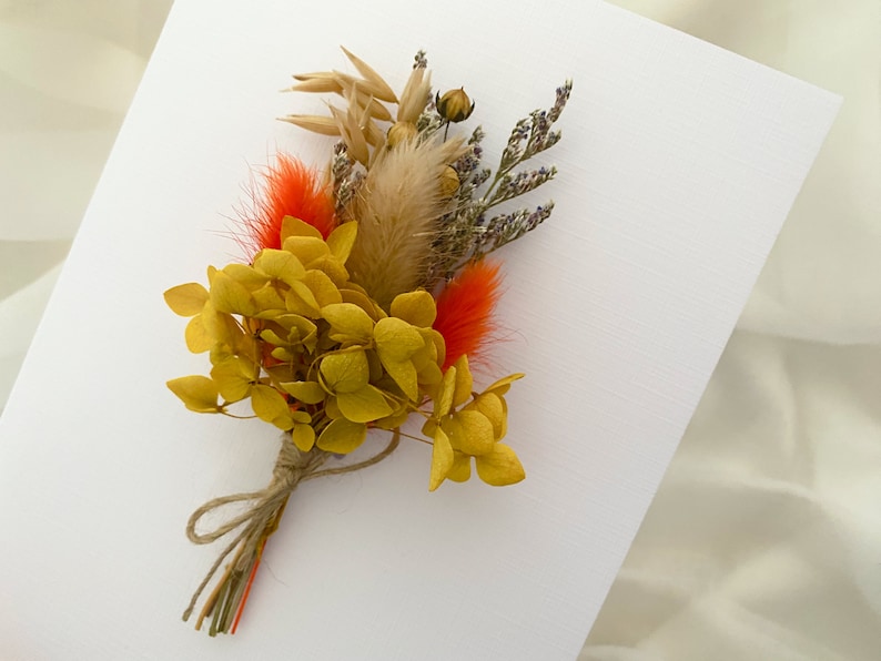 May include: A small bouquet of dried flowers, featuring yellow hydrangeas, orange feathers, and dried grasses, tied together with twine.