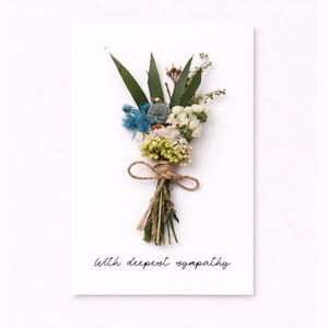 May include: A sympathy card with a small bouquet of dried flowers tied with twine. The bouquet includes green, blue, and white flowers and foliage. The words "With deepest sympathy" are written in a delicate script at the bottom of the card.