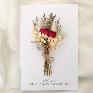 May include: A white card adorned with a dried flower bouquet. The bouquet features two red roses, cream gypsophila, and silvery-green foliage, bound with twine. The card reads "Add your personalised message here."