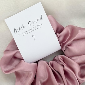 May include: A pink satin scrunchie with a white card that says "Bride Squad to have and to hold your hair back".