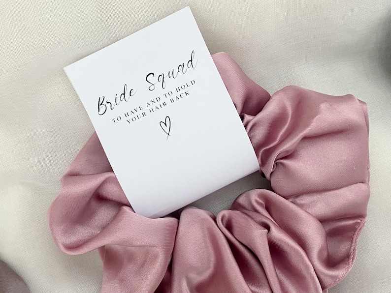 May include: A pink satin scrunchie with a white card that says "Bride Squad to have and to hold your hair back".