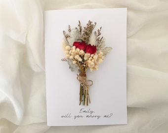 May include: A white card with a dried flower bouquet and the text "Emily, will you marry me?"