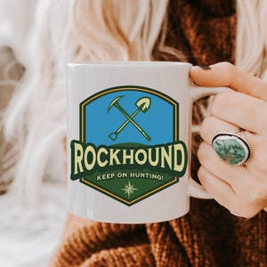 May include: White ceramic mug featuring a Rockhound design. The design includes a blue and green shield with crossed pickaxe and shovel, the word "ROCKHOUND", and the text "KEEP ON HUNTING!"