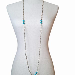 May include: A long necklace featuring a delicate silver-toned chain interspersed with small, off-white oval beads and vibrant turquoise-colored round beads. The chain hangs in a simple loop, showcasing a repeating pattern of beads throughout the length.