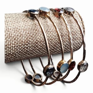 May include: Four thin, hammered metal bangles, each featuring a single round bezel-set gemstone. The stones include a dark blue, a pale blue, a deep red, and a clear stone with dark inclusions, all displayed on a cylindrical beige fabric stand.