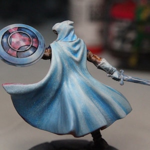 Puede incluir: Una figura en miniatura de un guerrero que lleva una capa azul fluida y sostiene una espada y un escudo con un diseño rojo y azul.