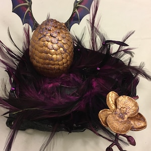 Puede incluir: Un huevo de dragón dorado con escamas moradas se encuentra en un nido de plumas moradas oscuras con una orquídea dorada a un lado.