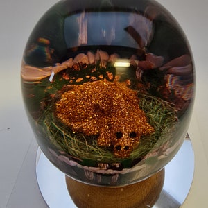 Puede incluir: Un pisapapeles de vidrio con una figurita de animal dorada brillante en su interior. La figurita está rodeada de hierba verde y flores rosas. El pisapapeles está sobre una base de madera.