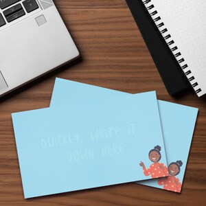 Op de afbeelding: Lichtblauwe notecards met de tekst "QUICKLY, WRITE IT DOWN HERE" in het wit. De kaarten bevatten illustraties van twee meisjes in oranje polkadotshirts. De kaarten liggen op een houten oppervlak, met een laptop en een notitieboek op de achtergrond.