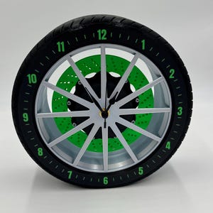 May include: A black and silver clock shaped like a car tire with green numbers and a green brake rotor design.