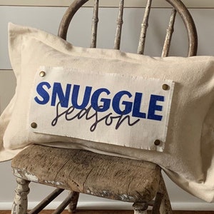May include: A white pillow with the words "Snuggle Season" in blue lettering on a distressed wooden chair.