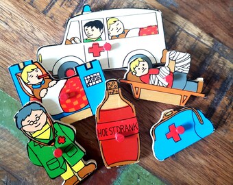 May include: A wooden puzzle set with a white ambulance, a doctor, a patient in bed, a patient on a stretcher, a bottle of cough syrup labeled "HOESTDRANK", and a blue first aid kit with a red cross.