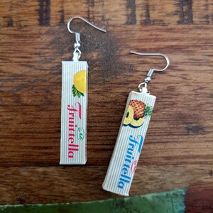 May include: A pair of silver dangle earrings with a rectangular charm featuring a vintage Fruittella candy box design. One charm has a pineapple design and the other has a lemon design.