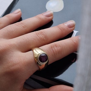 May include: A gold ring with a large, oval, dark red gemstone set in a bezel. The ring has a smooth, polished band and a classic, elegant design. The ring is shown on a person's finger.