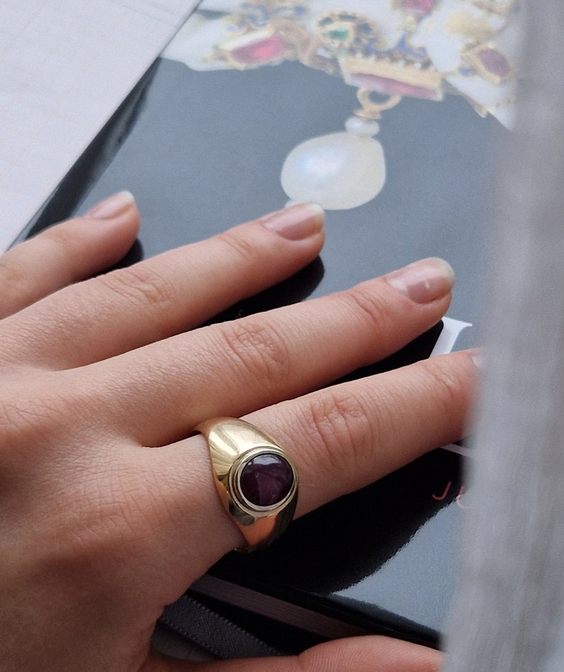 May include: A gold ring with a large, oval, dark red gemstone set in a bezel. The ring has a smooth, polished band and a classic, elegant design. The ring is shown on a person's finger.