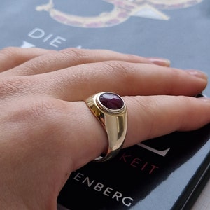 May include: A gold ring with a large oval ruby gemstone. The ring has a polished finish and a classic design. The ruby is a deep red color and is set in a bezel setting. The ring is on a person's finger.
