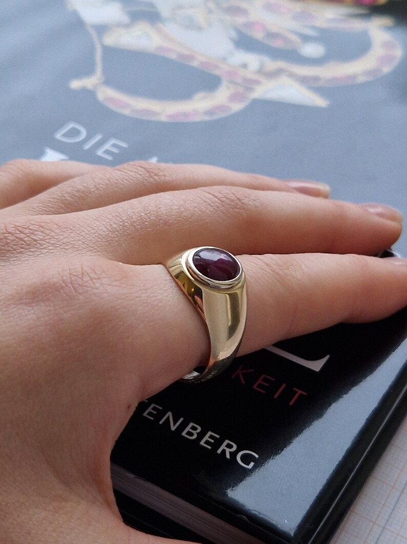 May include: A gold ring with a large oval ruby gemstone. The ring has a polished finish and a classic design. The ruby is a deep red color and is set in a bezel setting. The ring is on a person's finger.