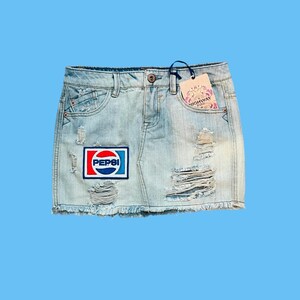 May include: Light blue denim skirt with distressed details and a frayed hem. Features a Pepsi logo patch and a tag that reads "HIGHWAY KIDS". The skirt has a classic five-pocket design.