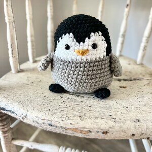 May include: A crocheted penguin toy with black and white fur, a yellow beak, and black eyes. The penguin is sitting on a white wooden chair.