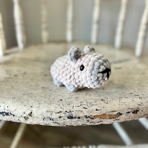May include: A small, gray crocheted mouse toy with black eyes. The toy is sitting on a white, distressed wooden chair.