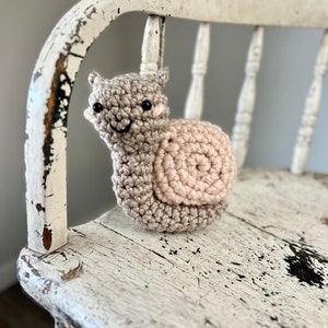 May include: A crocheted snail toy with a pink shell and a beige body. The snail has black eyes and a smiling mouth. The toy is sitting on a white wooden chair.