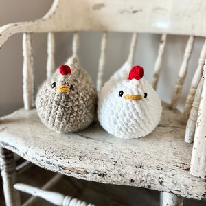 May include: Two crocheted chicken plushies, one white and one brown, sitting on a white wooden chair. The chickens have red combs and yellow beaks.