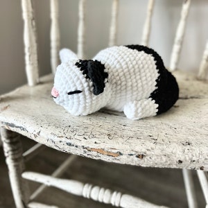 May include: A crocheted black and white cat toy, lying on a white wooden chair. The cat has a pink nose and black eyes.