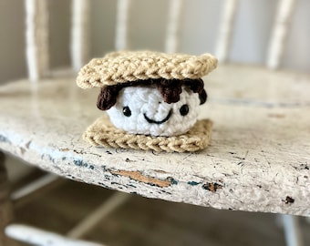 Könnte beinhalten: Ein gehäkeltes S'more mit einer weißen Marshmallow, brauner Schokolade und braunen Graham-Crackern. Das S'more hat ein lächelndes Gesicht.