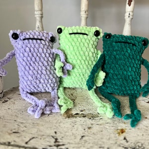 May include: Three crocheted frog plushies in different colors: lavender, light green, and dark green. The frogs are sitting on a white wooden chair.