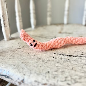 May include: A pink crocheted worm with black eyes, lying on a white wooden chair.