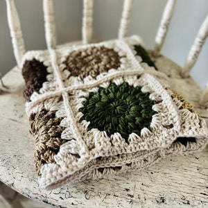 May include: A crocheted blanket with a square pattern in shades of brown, green, and cream. The blanket is folded and placed on a white wooden chair.