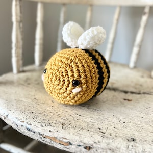 May include: A crocheted bee toy with a yellow and black striped body, white wings, and black eyes. The bee is sitting on a white wooden chair.