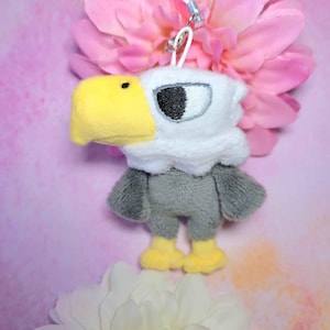 Peut inclure: Une peluche en forme d'aigle avec une tête blanche, un bec jaune et un corps gris. L'aigle a un œil noir et blanc et des pieds jaunes. Il est attaché à une ficelle blanche avec un fermoir en métal. Le fond présente des fleurs roses et blanches.