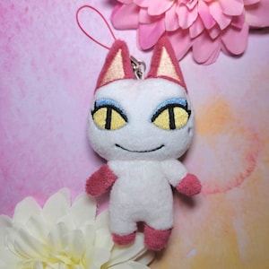 Peut inclure: Un chat en peluche blanc avec des accents roses sur les oreilles, les pattes et les pieds. Le chat a des yeux jaunes avec des pupilles noires et un eye-liner bleu. Une boucle rouge est attachée en haut. Fleurs roses et blanches en arrière-plan.