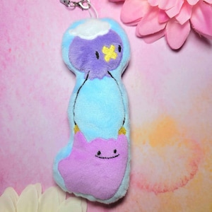 Può includere: Un portachiavi in peluche con un personaggio dei cartoni animati blu e viola con un dettaglio giallo. Il portachiavi ha un piccolo cinturino con una chiusura in metallo. Lo sfondo è una sfumatura rosa e arancione. Il peluche è morbido.