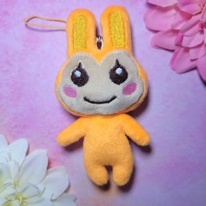 Puede incluir: Un llavero de peluche de conejo naranja con una cara sonriente, ojos marrones y mejillas rosadas. El conejo tiene orejas amarillas y lleva un atuendo naranja claro.