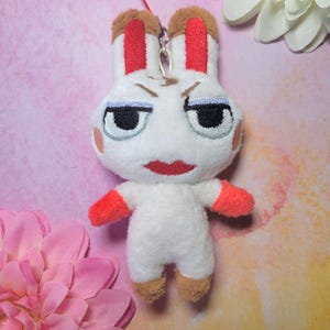May include: A white plush rabbit with a stern expression, red accents on the ears and arms, and brown feet. The rabbit has black eyes, a red mouth, and blush marks on its cheeks. A red string is attached to the top for hanging.