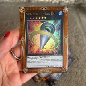 May include: A trading card featuring the text "NUMBER 11: BIG EYE" with a gold border. The card has a central design of a silver and gold shape with a green and blue background. The card's details include "SPELLCASTER/XYZ/EFFECT" and "ATK/2600 DEF/2000".