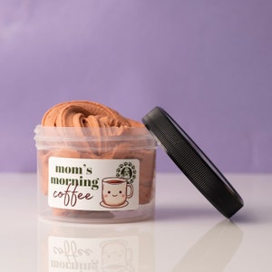May include: A clear plastic jar filled with brown-colored slime, with the lid slightly ajar. The jar has a label that reads "mom's morning coffee" with a smiling coffee cup graphic. The background is a solid purple color.