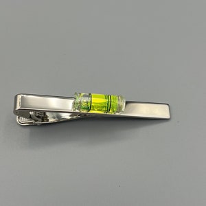 Può includere: Un fermaglio per cravatta argentato con un piccolo livello attaccato alla parte superiore. Il livello è verde e giallo e ha una bolla al centro.
