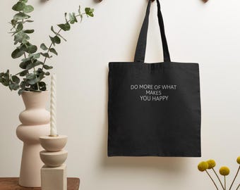 May include: Black tote bag with white text that reads "Do more of what makes you happy". The bag is hanging on a wooden wall rack.