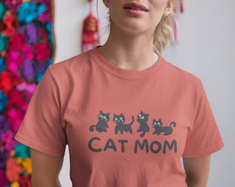 May include: A woman wearing a coral colored t-shirt with a graphic of four black cats and the text "CAT MOM".