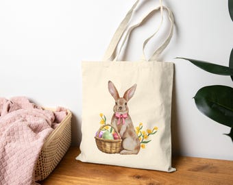 Peut inclure: Un sac fourre-tout beige avec un lapin brun tenant un panier d'œufs de Pâques colorés. Le lapin porte un nœud rose. Le sac a des fleurs jaunes sur le côté.