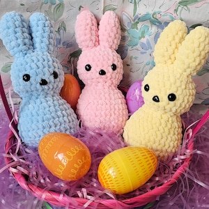 May include: Three crocheted bunny plushes in a pink basket with purple shredded paper. The bunnies are light blue, pink, and yellow. There are also orange and yellow Easter eggs in the basket.