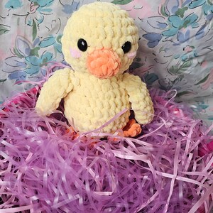 Puede incluir: Un pollito amarillo tejido a mano con ojos negros, mejillas rosas y pico y patas naranjas. El pollito está sentado en una cesta rosa llena de papel triturado morado. El fondo es un estampado floral.