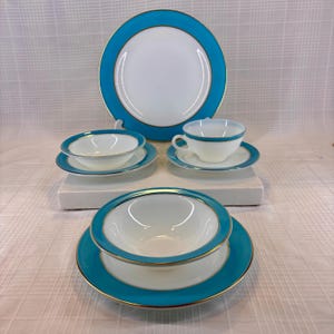 May include: A set of white and turquoise dishware with gold trim. The set includes a large plate, bowls, saucers, and a teacup and saucer. The turquoise color is a vibrant accent against the white, creating an elegant look.